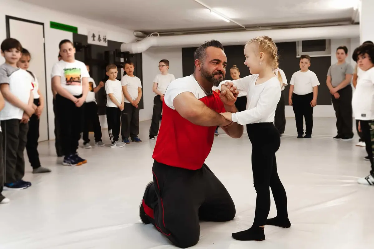 Master Kaan Firat kniet und übt mit einem Mädchen vor der Kindergruppe